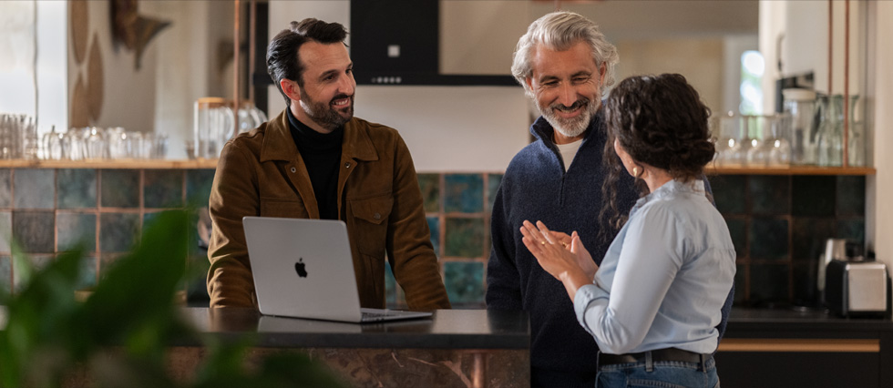 Dos hombres y una mujer están conversando, y se ve un MacBook sobre una mesa.