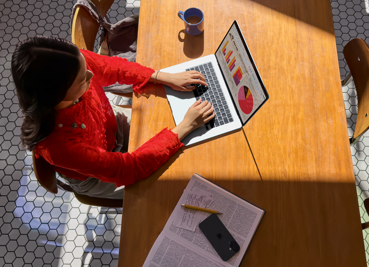 En person bruger MacBook ved et bord med et krus, en smartphone og papirer ved siden af.