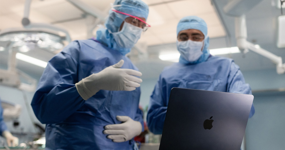 Dois cirurgiões olham para um MacBook na sala de cirurgia, e um gesticula com a mão enquanto o outro observa