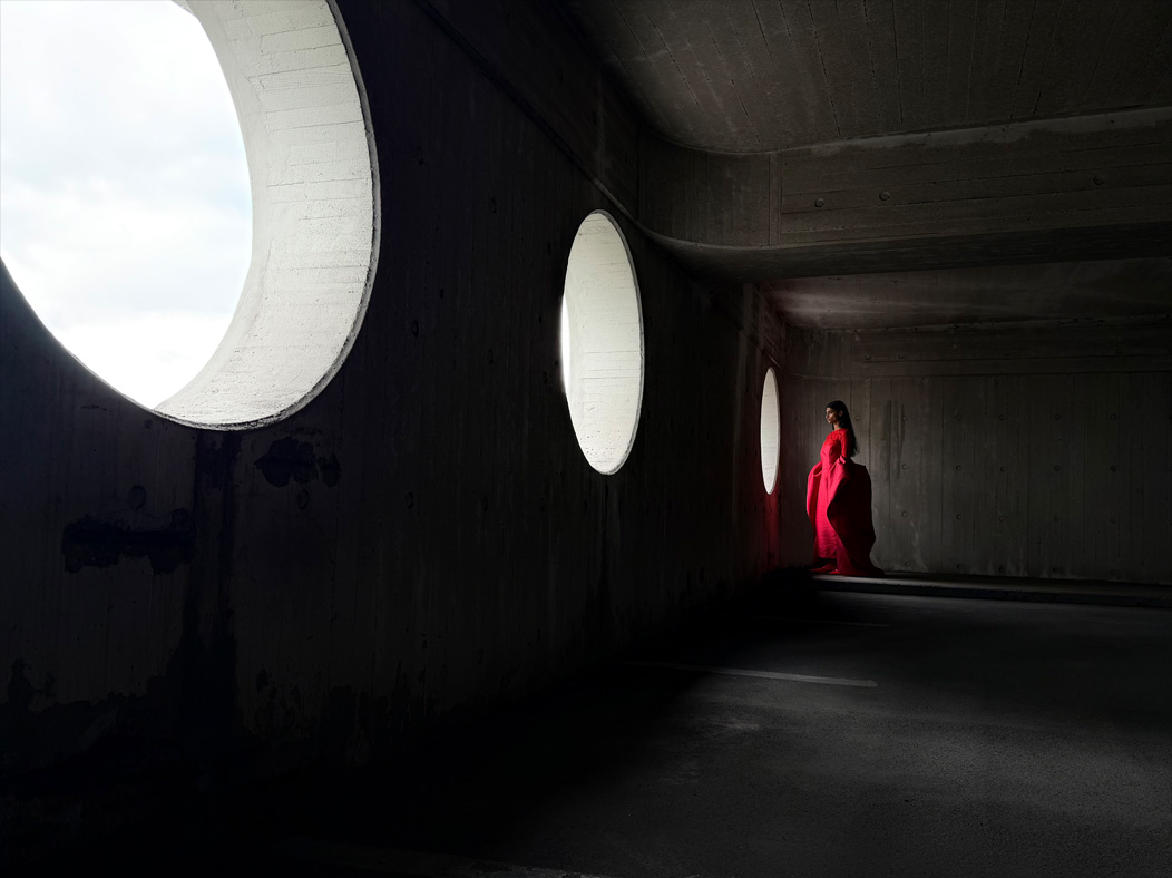 Une femme vêtue d’une longue robe rouge se fait photographier dans une pièce vide. L’app Appareil photo change de distance focale pour illustrer les capacités du système caméra pro, mettant en valeur : 28 mm